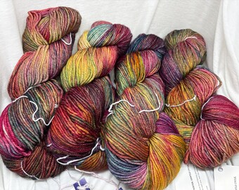 May include: A collection of five skeins of hand-dyed yarn featuring a variegated color palette of deep magenta, golden yellow, forest green, and hints of blue. Each skein is twisted and secured with a white string, showcasing a soft, plied texture.