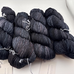May include: Four skeins of hand-dyed yarn in a dark charcoal and black colorway with subtle grey marbling throughout. Each skein is twisted and features a white paper tag attached with a string, displaying printed text and care instructions.
