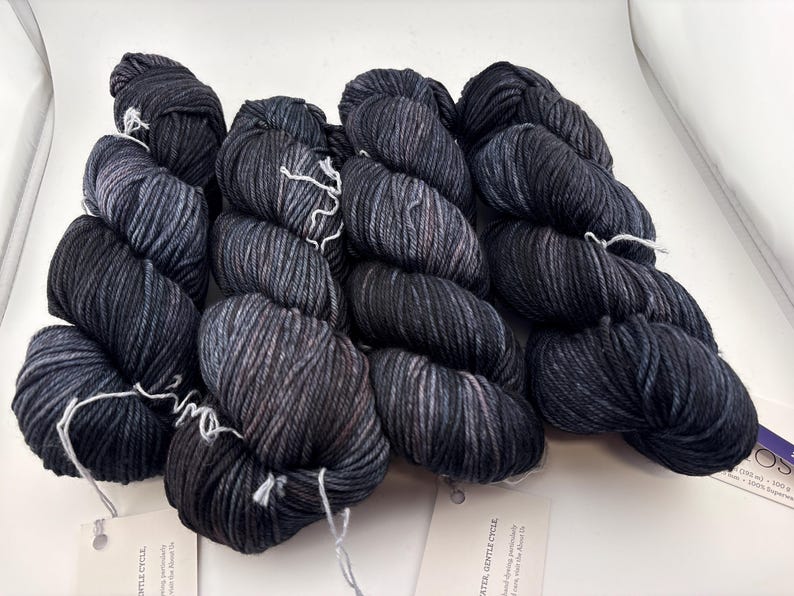 May include: Four skeins of hand-dyed yarn in a dark charcoal and black colorway with subtle grey marbling throughout. Each skein is twisted and features a white paper tag attached with a string, displaying printed text and care instructions.