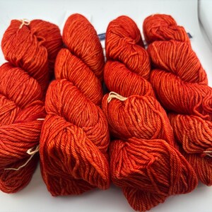 May include: Four skeins of vibrant orange yarn arranged side by side. Each skein is tightly twisted and features a consistent, saturated red-orange hue with a visible ply structure. The yarn is presented in a clean, uniform, and organized manner.