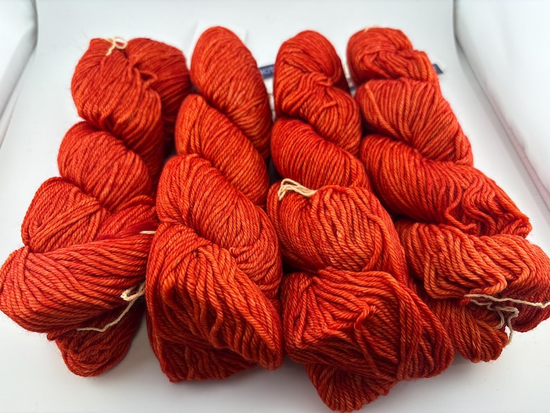 May include: Four skeins of vibrant orange yarn arranged side by side. Each skein is tightly twisted and features a consistent, saturated red-orange hue with a visible ply structure. The yarn is presented in a clean, uniform, and organized manner.