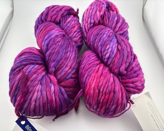 Puede incluir: Dos madejas de hilo grueso y voluminoso con una paleta de colores variados en magenta vibrante, morado intenso y rosa brillante. Cada madeja está retorcida en un paquete suave, con una pequeña etiqueta blanca de producto en la parte inferior.