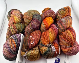 May include: Four skeins of hand-dyed yarn arranged in a square formation. The yarn features a variegated color palette of deep orange, golden yellow, olive green, and muted purple tones, with white paper labels attached to the bottom.