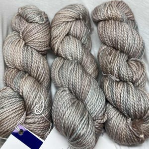 May include: Six skeins of yarn in a neutral color palette of gray, brown, and hints of blue. The yarn is tightly wound, showing a textured surface. A small tag is attached to one of the skeins. Ideal for knitting or crochet projects.
