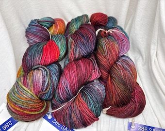 May include: Three skeins of hand-dyed yarn featuring a variegated color palette of deep red, magenta, forest green, and hints of golden yellow. Each skein is secured with a white string and includes a blue tag with the text 286 SAO.