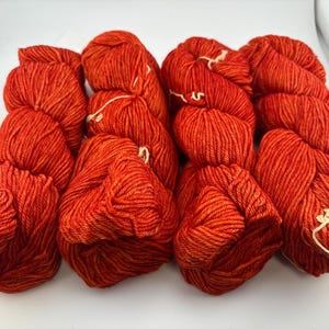 May include: Four skeins of vibrant orange yarn arranged in a row. Each skein features a twisted, multi-ply construction with a consistent, saturated color throughout. The yarn has a slight sheen and a textured appearance, with small white ties.