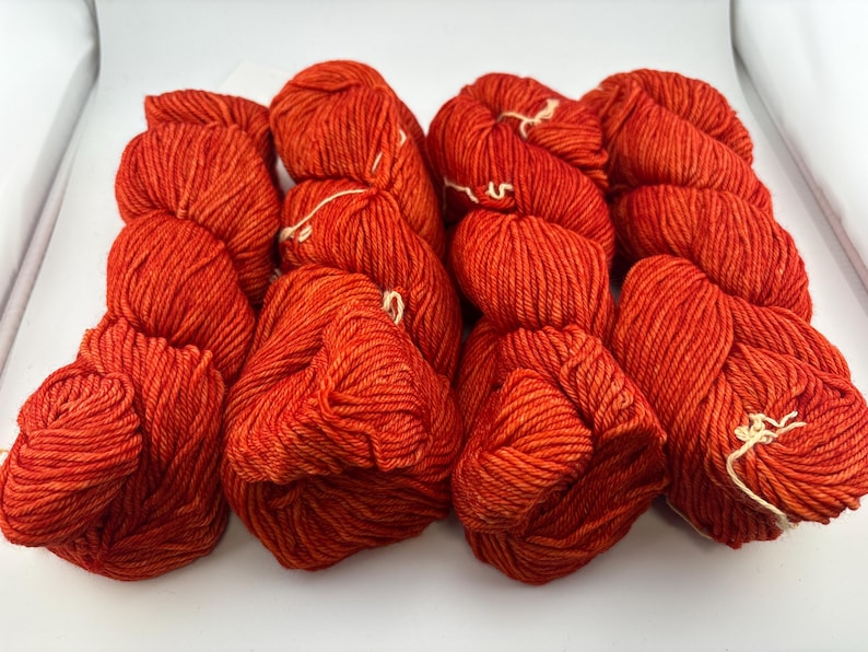 May include: Four skeins of vibrant orange yarn arranged in a row. Each skein features a twisted, multi-ply construction with a consistent, saturated color throughout. The yarn has a slight sheen and a textured appearance, with small white ties.