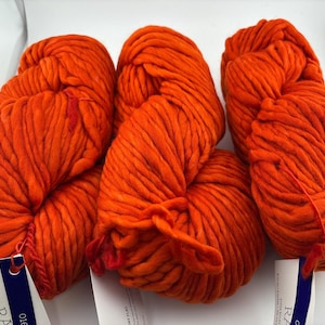 K&ouml;nnte beinhalten: Drei Str&auml;nge aus leuchtend orangefarbenem, super volumin&ouml;sem Garn. Jeder Strang hat eine dicke, gedrehte Textur und ist mit einem wei&szlig;en Papieretikett mit dunkelblauem Text versehen, einschlie&szlig;lich des Wortes RA, das unten sichtbar ist.