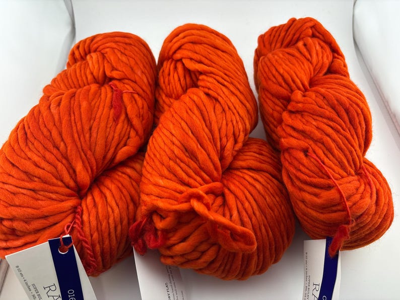 K&ouml;nnte beinhalten: Drei Str&auml;nge aus leuchtend orangefarbenem, super volumin&ouml;sem Garn. Jeder Strang hat eine dicke, gedrehte Textur und ist mit einem wei&szlig;en Papieretikett mit dunkelblauem Text versehen, einschlie&szlig;lich des Wortes RA, das unten sichtbar ist.