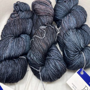 May include: Three skeins of hand-dyed yarn in a dark charcoal and navy blue colorway with subtle brown undertones. Each skein features a twisted texture and is secured with a white string. A blue and white product tag is attached to the yarn.