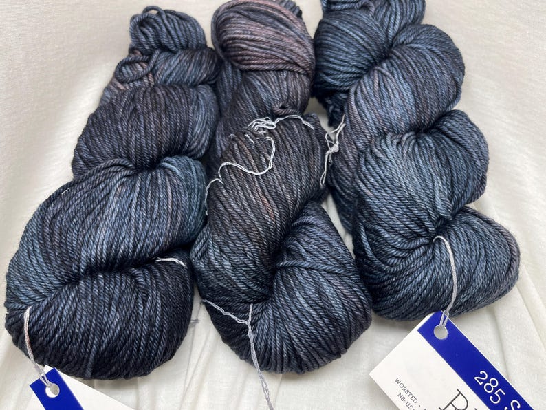 May include: Three skeins of hand-dyed yarn in a dark charcoal and navy blue colorway with subtle brown undertones. Each skein features a twisted texture and is secured with a white string. A blue and white product tag is attached to the yarn.