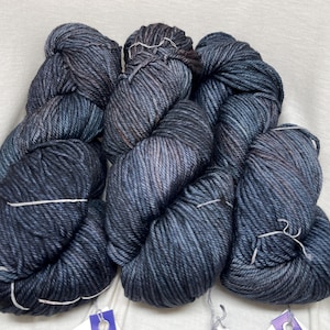 May include: Three skeins of hand-dyed yarn featuring a deep charcoal and navy blue color palette with subtle hints of brown. The yarn has a thick, twisted texture and is bundled into individual hanks, each secured with a small white string.