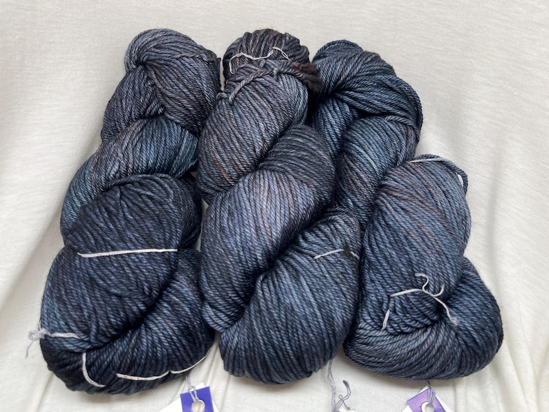 May include: Three skeins of hand-dyed yarn featuring a deep charcoal and navy blue color palette with subtle hints of brown. The yarn has a thick, twisted texture and is bundled into individual hanks, each secured with a small white string.