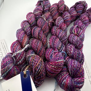 May include: Four skeins of hand-dyed yarn in a variegated colorway featuring deep magenta, purple, blue, and yellow speckles. Each skein is attached to a white paper label with the text 005 ANIVERSARIO, DK/SPORT, 160 yd, and 100% Superwash Merino Wool.