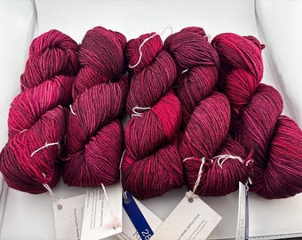 Könnte beinhalten: Fünf Stränge aus tief magenta- und schwarzfarbenem, meliertem Garn, die in einer Reihe angeordnet sind. Jeder Strang hat eine gedrehte Textur und ist mit einem weißen Papieretikett versehen, das Pflegehinweise wie cold water und gentle cycle enthält.