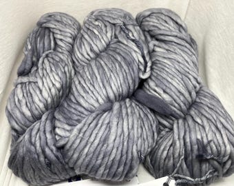 Puede incluir: Primer plano de varios ovillos de hilo grueso gris. El hilo parece ser lana merino súper gruesa, con un aspecto suave y texturizado. El color es una mezcla de gris claro y oscuro, creando un efecto veteado. Ideal para proyectos de tejido o ganchillo.
