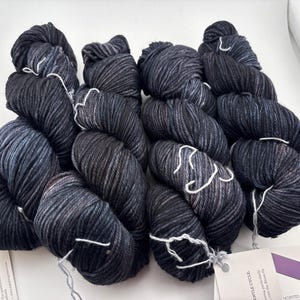 May include: Four skeins of worsted weight yarn in a variegated dark charcoal and black colorway with subtle blue undertones. Each skein features a twisted texture and is secured with a white paper label containing printed product information.