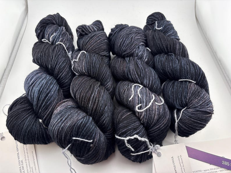 May include: Four skeins of worsted weight yarn in a variegated dark charcoal and black colorway with subtle blue undertones. Each skein features a twisted texture and is secured with a white paper label containing printed product information.