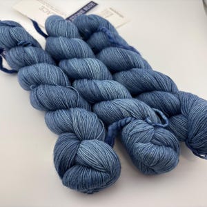 May include: Three skeins of yarn in a variegated stone blue colorway. Each skein is twisted into a compact bundle and features a white paper label attached to the top. The yarn displays a subtle tonal variation with lighter and darker blue fibers.