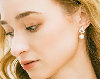 May include: A woman wearing a white dress with a gold necklace and pearl earrings. The necklace features a pearl pendant with a small floral design.