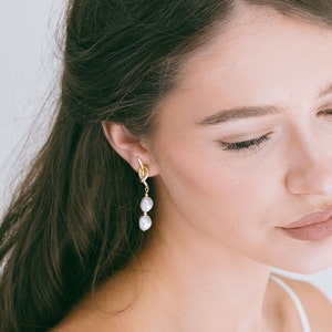Peut inclure: Une femme aux longs cheveux bruns portant un débardeur blanc et des boucles d'oreilles dorées avec des accents de perles.