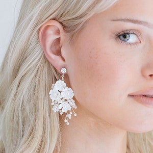 May include: A woman with blonde hair wearing white floral earrings. The earrings are made of white flowers and have a silver finish.