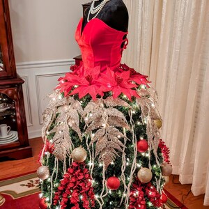 Op de afbeelding: Een kerstboom versierd met rode en gouden kerstballen, witte parelkransen en rode kerststerren. De boom is gevormd als een jurk en wordt getoond op een mannequin.
