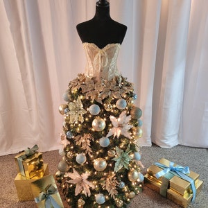 Op de afbeelding: Een kerstboom versierd met gouden en blauwe ornamenten, lichtjes en een wit en gouden korset bovenop. De boom is omringd door gouden cadeaudozen met blauw lint.