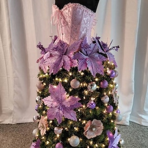 Op de afbeelding: Een roze satijnen corset wordt gebruikt als basis voor een kerstboom die is versierd met paarse kerststerren en zilveren en paarse kerstballen.
