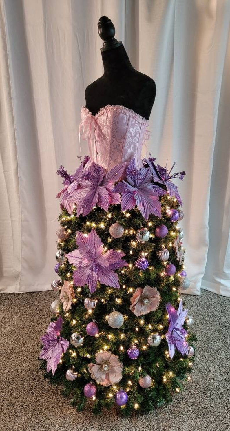 Op de afbeelding: Een roze satijnen corset wordt gebruikt als basis voor een kerstboom die is versierd met paarse kerststerren en zilveren en paarse kerstballen.