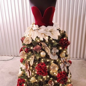 Op de afbeelding: Een kerstboom versierd met rode en gouden ornamenten, witte kerststerren en witte veren. De boom is gevormd als een jurk en staat op een zwart mannequin.