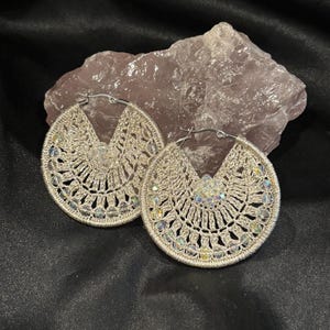 May include: A pair of silver-toned, crocheted hoop earrings. The earrings feature a circular design with intricate openwork patterns and are adorned with iridescent beads. The earrings are displayed on a black surface with a pinkish-gray rock in the background.