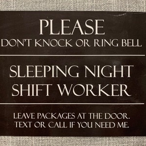 May include: A black rectangular sign with white text. The sign reads "PLEASE DON'T KNOCK OR RING BELL SLEEPING NIGHT SHIFT WORKER." Additional text at the bottom says "LEAVE PACKAGES AT THE DOOR. TEXT OR CALL IF YOU NEED ME."