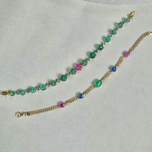 May include: Two gold bracelets with colorful gemstones. One bracelet features green, pink, and blue stones, while the other has green, pink, and blue gemstones. Both bracelets have gold clasps.