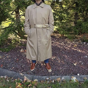 May include: A beige trench coat featuring a classic collar, long sleeves, and a matching fabric belt with a metal buckle at the waist. The coat has a button-front closure, two side pockets, and a mid-calf length hemline.