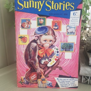 A vintage children's activity book cover featuring a cartoon monkey painting with a paintbrush and a palette. The cover is pink with a blue title "Sunny Stories" and a price of 6d. The text below the monkey says "Do you like my paintings? To find what I am write the first letter of each object."