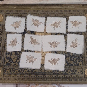 Może przedstawiać: Dziesięć małych białych kwadratów z tkaniny z nadrukowanym na każdym brązowym wzorem pszczoły. Kwadraty mają strzępiony brzeg.