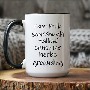 Op de afbeelding: Witte keramische mok met een zwarte handgreep en rand. De mok heeft de woorden "raw milk, sourdough, tallow, sunshine, herbs, grounding" in zwart schrift. De mok staat op een houten oppervlak.