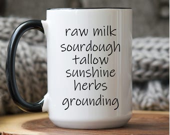 Könnte beinhalten: Weißer Keramikbecher mit schwarzem Henkel und Rand. Der Becher trägt die Worte "raw milk, sourdough, tallow, sunshine, herbs, grounding" in schwarzer Schrift. Der Becher steht auf einer Holzoberfläche.