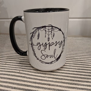 May include: White ceramic mug with a black rim and handle. The mug features a black and white design of a dreamcatcher with feathers and the text "gypsy soul".