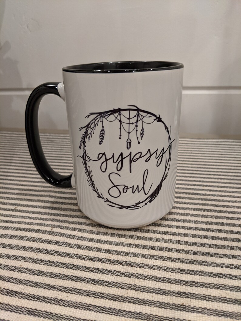 May include: White ceramic mug with a black rim and handle. The mug features a black and white design of a dreamcatcher with feathers and the text "gypsy soul".