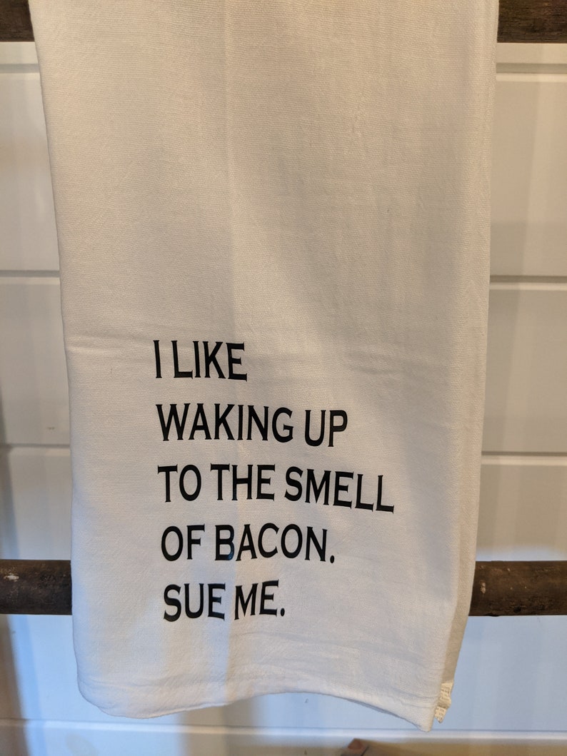 Op de afbeelding: Witte keukendoek met zwarte tekst die luidt: "Ik vind het leuk om wakker te worden met de geur van bacon. Dagvaard me."