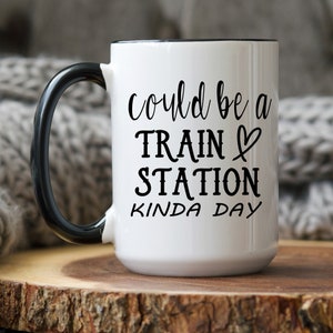 May include: White ceramic coffee mug with a black rim and handle. The mug has black text that reads "could be a train station kinda day".