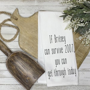 May include: White kitchen towel with black text that reads "If Britney can survive 2007, you can get through today".