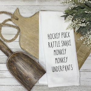 May include: White kitchen towel with black text that reads "Hockey Puck, Rattle Snake, Monkey, Monkey, Underpants". The towel is lying on a wooden surface with a wooden cutting board and a wooden scoop.