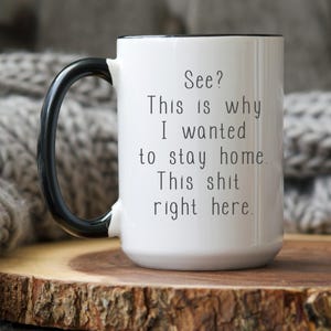 Op de afbeelding: Witte keramische mok met een zwarte handgreep en rand. De mok heeft de tekst "See? This is why I wanted to stay home. This shit right here." in het zwart. De mok staat op een houten oppervlak.