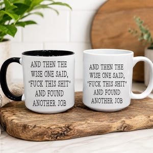 Op de afbeelding: Twee keramische mokken met zwarte tekst: AND THEN THE WISE ONE SAID, "FUCK THIS SHIT" AND FOUND ANOTHER JOB. Eén mok heeft een zwart handvat en een zwarte binnenkant, terwijl de andere volledig wit is. Beide mokken hebben een cilindrische vorm.