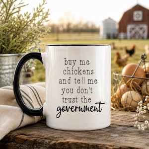 Op de afbeelding: Een witte keramische mok met een zwarte handgreep en binnenrand. Op de voorzijde van de mok staat zwarte tekst: buy me chickens and tell me you don't trust the government. De mok heeft een gladde afwerking en een standaard cilindrische vorm.