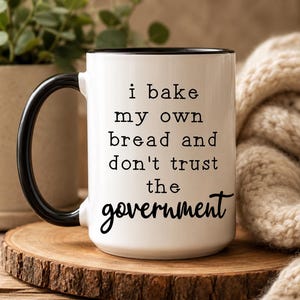 May include: A white ceramic mug with a black handle and black interior rim. The mug features black text that reads: i bake my own bread and don't trust the government. The font for the first four lines is a simple typewriter style, while the word government is in a cursive script.