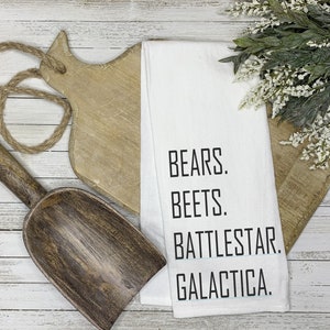 Op de afbeelding: Witte keukendoek met zwarte tekst die "Bears. Beets. Battlestar. Galactica." luidt.